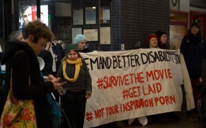 Demonstrators outside the Embassy Theatre Wellington. The banner they are holding reads "Demand better stories #Survivethemovie #GetLaid #NotYourInspirationPorn. A male and female figure are silhouetted together in black. Photo by Zoë Deans